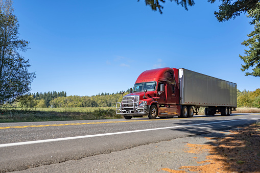 Classic big rig red bonnet semi truck with high roof transporting commercial cargo in dry van semi trailer running for delivery on the winding highway road with summer green trees on the side