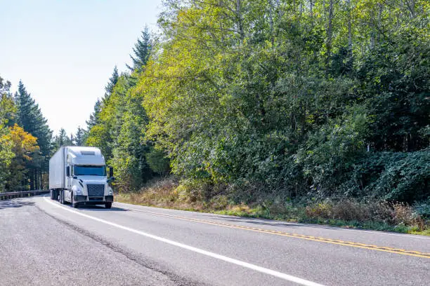 Photo of White comfort big rig semi truck transporting cargo in refrigerator semi trailer running on the turning road with green forest on the sides