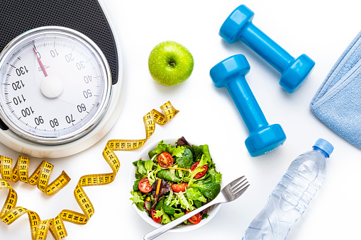 Top view of a healthy salad and exercising background concept: The composition includes a fresh salad, a water bottle, a towel, two dumbbells, a tape mesure and a weight scale on white background.