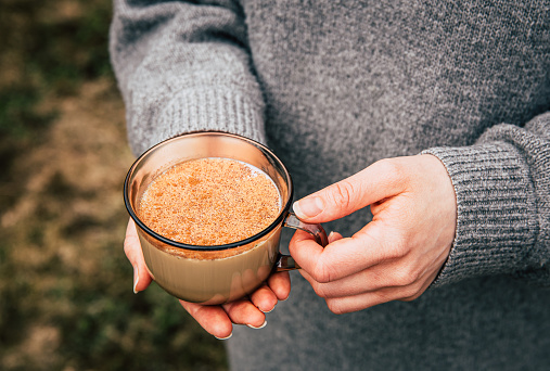 Close up view of woman hands wearing gray knitter cozy sweater holding cup of warm Chai latte with cinnamon powder outdoors in autumn. Picnic outdoors concept.