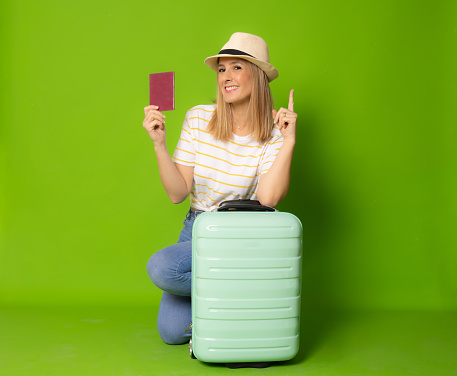 Young smiling woman with suitcase holding passport pointing finger up isolated over green background.