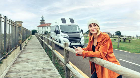 Young woman looking camera leaning on fence in front of camper van with lighthouse in background