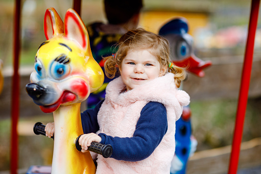 Adorable little toddler girl riding on animal on roundabout carousel in amusement park. Happy healthy baby child having fun outdoors on sunny day. Family weedend or vacations.