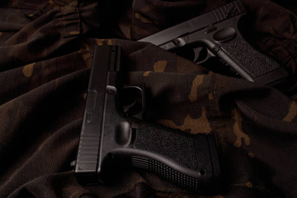 istock Two black pistols are lying on a camouflage soldier's jacket. A firearm that makes you think about self-defense. World Arms Day. Side view from above 1351298407