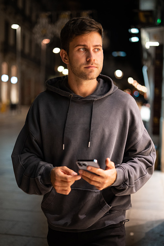 Man in casual hoodie using smartphone and looking away while walking on city street at night