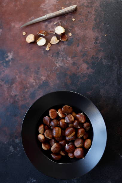 Chestnuts in a black deep plate on a rough concrete floor with a black-brown rust effect. At the top of the photo are chestnuts and their shells peeled with a wooden-handled knife.