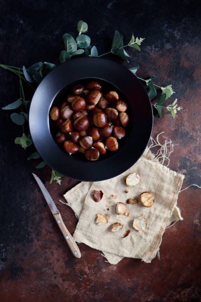 istock Chestnuts in a black deep plate on a rough concrete floor with a black-brown rust effect. Next to the plate are peeled chestnuts and their shells on burlap with a wooden-handled knife. 1351301084