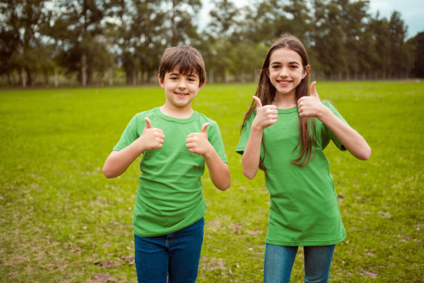 Brother and sister during environmental cleanup. They are at park. Smiling environmentalists wearing green t-shirts, looking at camera with thumbs up.
