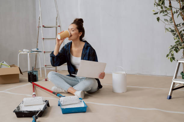 istock An architectural interior designer sits on the floor in an empty apartment, drinking takeaway coffee, holding laptop on lap, designing flat layout, around her painting equipment brushes rollers paints 1351313684