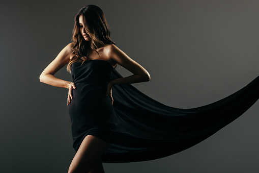 Beautiful young woman posing pregnant in studio. She is wrapped in long black textile that falls behind her. She has a long healthy hair and looks very sensual