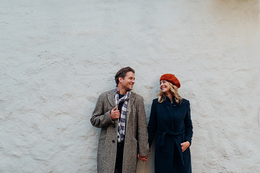 Smiling man and woman holding hands and standing over a white wall in the city