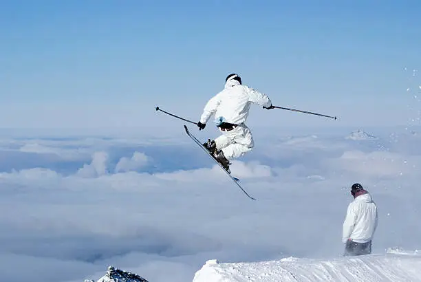 Extreme skiier in white jumping high above the clouds as a friend, also dressed in white, looks on.