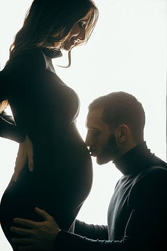 Beautiful couple posing in studio. She is pregnant in last trimester and looks amazing and man is kissing her abdomen. They both wear black clothes. Strong backlight