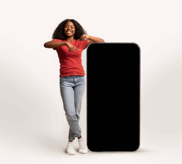 Cheerful Young African American Lady Leaning And Pointing At Big Blank Smartphone Cheerful Young African American Lady Leaning And Pointing At Big Smartphone With Blank Screen, Smiling Black Lady Showing Copy Space For Advertisement, Standing Over White Studio Background, Mockup leaning stock pictures, royalty-free photos & images