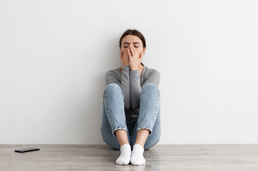 Despaired sad worried caucasian millennial female suffering from breakup, mental problems and bad news sitting on floor at home, on gray wall background. Panic attack, depression and stress alone