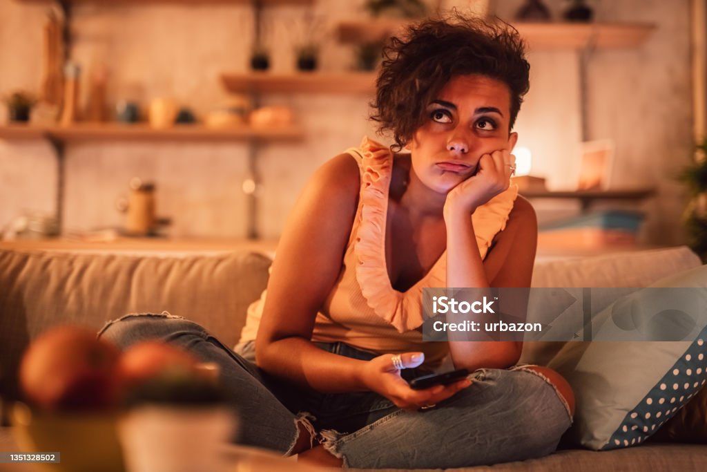 Bored woman sitting on sofa and holding phone Relationship problems, bad news, bored at home with modern gadget. Sad cute millennial woman looking at smartphone, sitting on couch alone in living room interior, indoors. Boredom Stock Photo