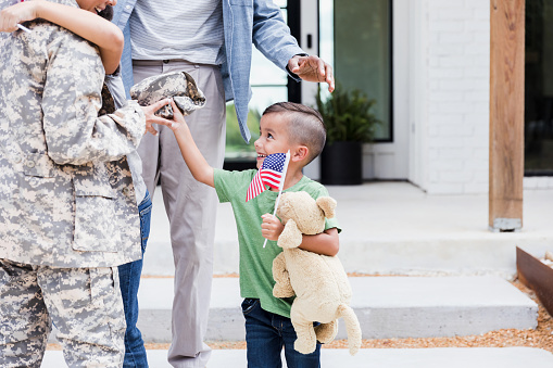 As his brother embraces their mother when she comes home, the elementary age boy holds his American flag and his toy bear as he reaches for the unrecognizable soldier mother's uniform cap.