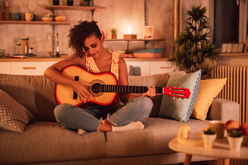 Young beautiful woman playing acoustic guitar on sofa at home in the evening.