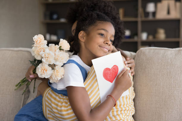 Dreamy african american girl cuddling loving mother. Adorable smiling dreamy pretty african american little kid girl cuddling happy young beautiful mother, presenting flowers bouquet and paper card with heart symbol, congratulating with birthday at home birthday wishes for daughter stock pictures, royalty-free photos & images