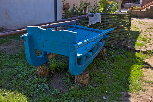 An old blue wooden sleigh stands on logs near a rural house.