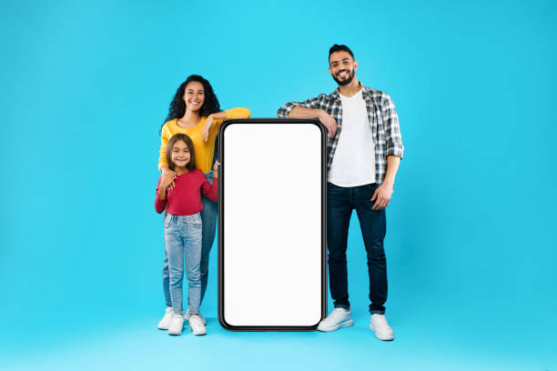 Arab Family Standing Near Huge Phone Screen On Blue Background Arab Family Standing Near Huge Phone With Empty Screen For Mobile Application Advertisement Posing On Blue Studio Background. Arabic Parents And Daughter Advertising App. Mockup middle eastern ethnicity photos stock pictures, royalty-free photos & images