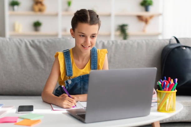 istock Cheerful girl sitting at desk, using laptop, drawing in copybook 1351335385