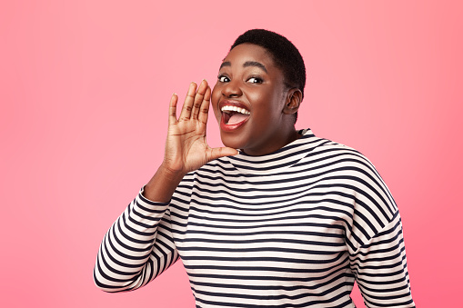 Mujer negra con sobrepeso gritando de la mano cerca de la boca, fondo rosado photo