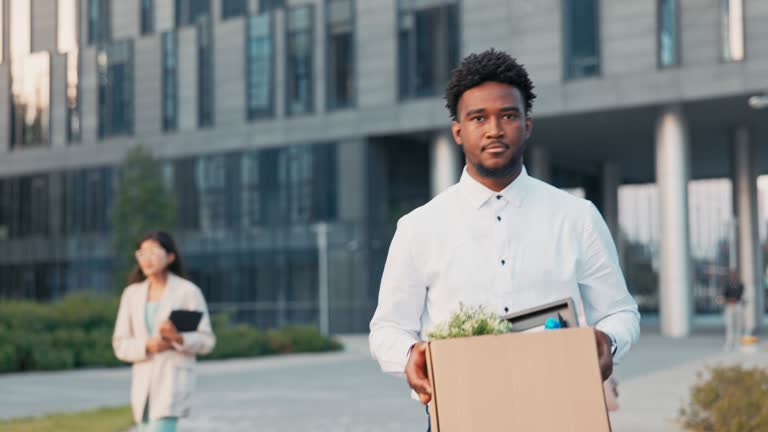 A dark-skinned man long-time office worker is fired from job, walks out of company building with co-workers, holding box of packed belongings in hands, a disgruntled expression, anger, sadness