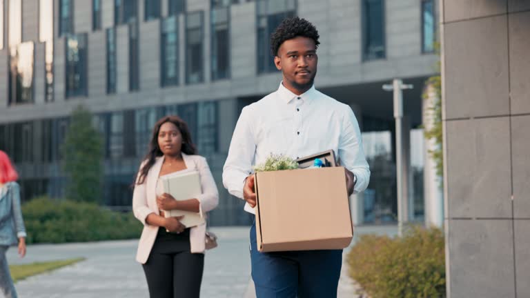 A dark-skinned man long-time office worker is fired from job, walks out of company building with co-workers, holding box of packed belongings in hands, a disgruntled expression, anger, sadness