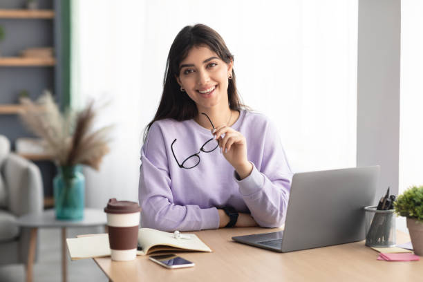 Cheerful millennial lady sitting at desk and posing with glasses Portrait Of Smiling Woman Sitting At Table With Notebook Holding Eyeglasses And Posing Looking At Camera. Student Studying Remotely, Psychologist Working Distantly On Laptop, Consulting Clients Online mental health professional stock pictures, royalty-free photos & images