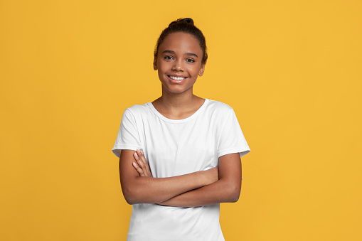 Happy calm teen black girl pupil in white t-shirt with crossed arms, isolated on yellow background, empty space. Positive schoolgirl, student emotions, good ad and covid-19 quarantine, studio shot