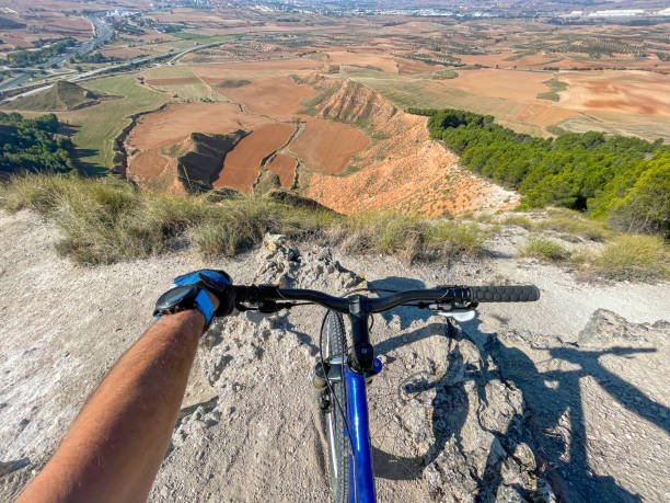 vista in prima persona di un ciclista su una mountain bike sul bordo di una scogliera su un sentiero di montagna. sullo sfondo campi coltivati e la periferia di una città - bikers point of view foto e immagini stock