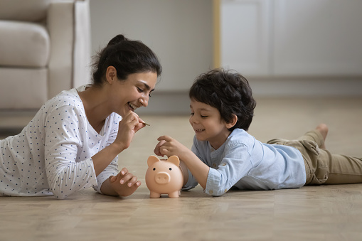 Caring young asian indian mother teaching little kid son saving money or planning future purchases, putting coins in piggybank, lying on heated floor, financial education for children concept.