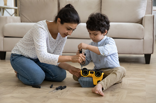 Happy young indian ethnic mother helping adorable small kid son fixing toy car with screwdriver, playing together at home. Smiling asian mum teaching little child repairing, developing motility skills