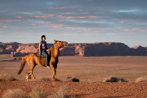 Pretty Teenage Navajo Native American Girl Riding her Horse with Dog in the Monument Valley Tribal Park on the Utah/Arizona border in the United States