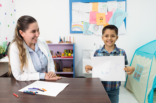 A happy little latin boy standing next to a female psychologist and showing his drawing to the camera with a smile.