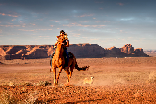 Pretty Teenage Navajo Native American Girl Riding her Horse with Dog in the Monument Valley Tribal Park on the Utah/Arizona border in the United States