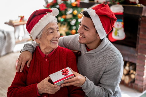 Cute grandson surprising his grandmother both looking at each other while he holds a christmas gift very lovingly - Celebration concepts