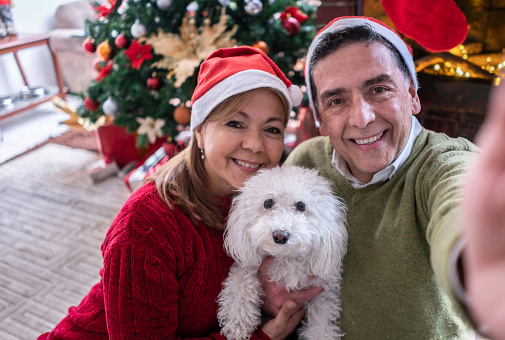 Latin American mature couple at home wearing Santa's hats and posing with their cute dog while taking a selfie on a beautiful christmas day - Christmas and lifestyles concepts
