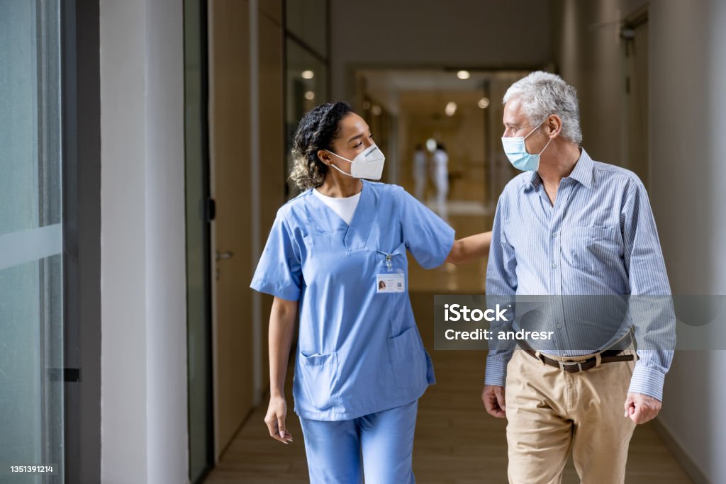 Doctor talking to a patient in the corridor of a hospital while wearing face masks African American doctor talking to a patient in the corridor of a hospital while wearing face masks during the COVID-19 pandemic. **DESIGN ON NAME TAG WAS MADE FROM SCRATCH BY US** Patient Stock Photo