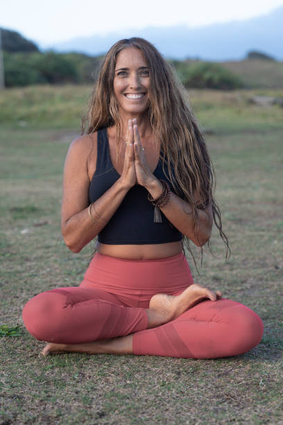 Yoga instructor on the grass near the beach, wearing a black top and red pants, in a Lotus Position, with hands in prayer position A  vertical shot of a Yoga instructor on the grass near the beach wearing a black top and red pants, smiling, in Lotus Position and hands in Prayer Position, there is grass in the foreground and background yoga instructor stock pictures, royalty-free photos & images