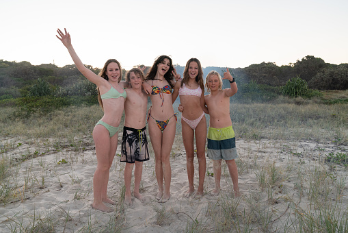 A group of teenagers  standing on the sand at the beach together, they are wearing bikinis and board shorts, there is a blue smoke bomb in front of them and grass and trees behind them