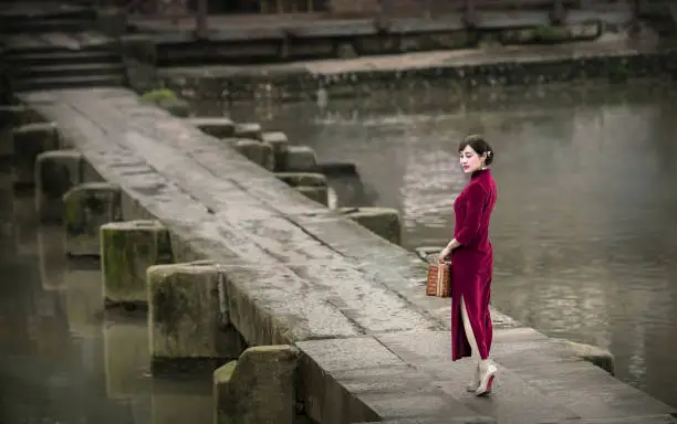A woman in a qipao carrying a suitcase walks across an ancient stone slab bridge