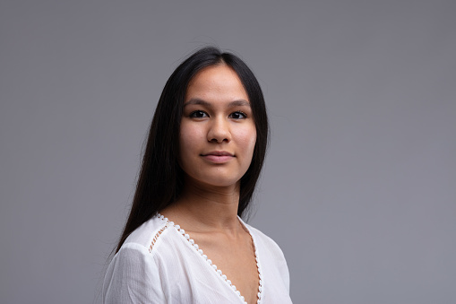 Portrait of a young woman with long straight dark hairlooking thoughtfully at the camera over a grey background