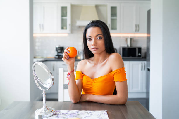 Beautiful young asian brunette sitting at the kitchen table with a mirror on it and holding an orange in her hand