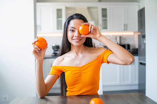 Beautiful and funny young brunette with orange shirt  holding two oranges in front of her face with toothy smile. Domestic background. Fun at home. Breakfast preparation