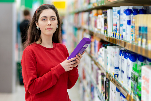 A young Caucasian brunette woman in a red sweatshirt is holding a bottle of shampoo. In the background, shelves with cosmetics. The concept of buying cosmetics.