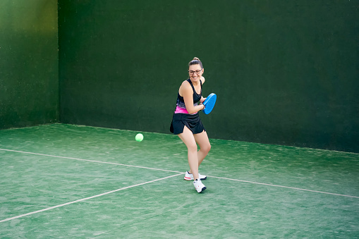 Female athlete hitting ball with racket while playing padel in spacious gym with green walls