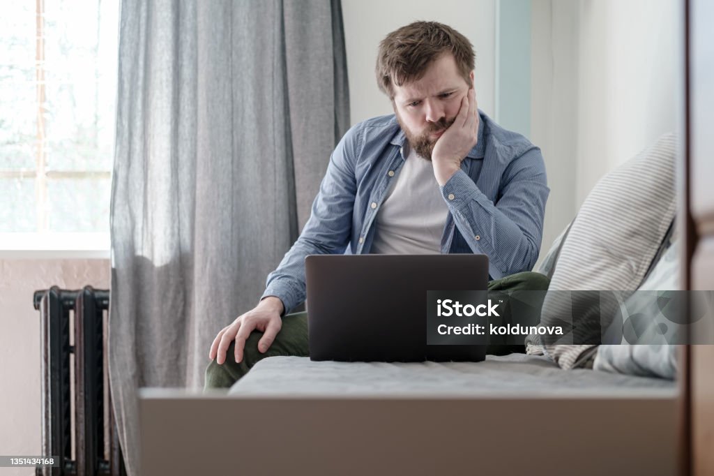 Man with a laptop is bored while sitting at home. He is sad because he is forced to stay home during a virus and pandemic epidemic. Man with a laptop is bored sitting on a sofa at home. He is sad because he is forced to stay home during a virus and pandemic epidemic. Laptop Stock Photo