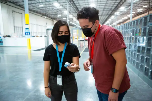 A woman using a handheld thermometer to take the temperature of a colleague at the entrance of a large fulfillment center.